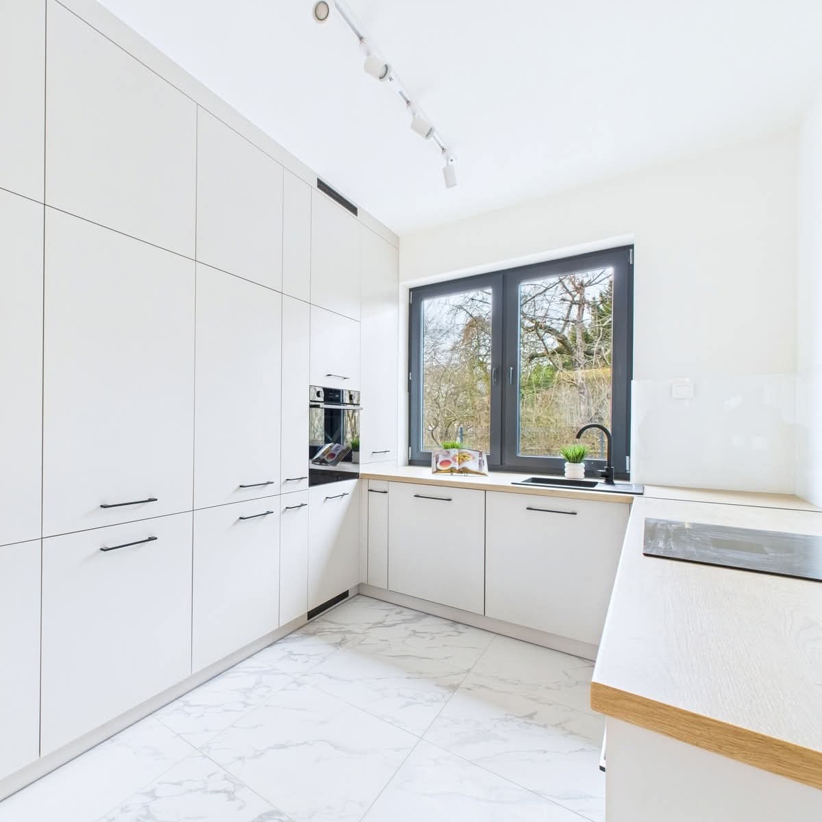 Biała kuchnia na wymiar z czarnymi akcentami: minimalistyczne fronty, marmurowe płytki podłogowe i okno z widokiem na zieleń. Nowoczesny design.