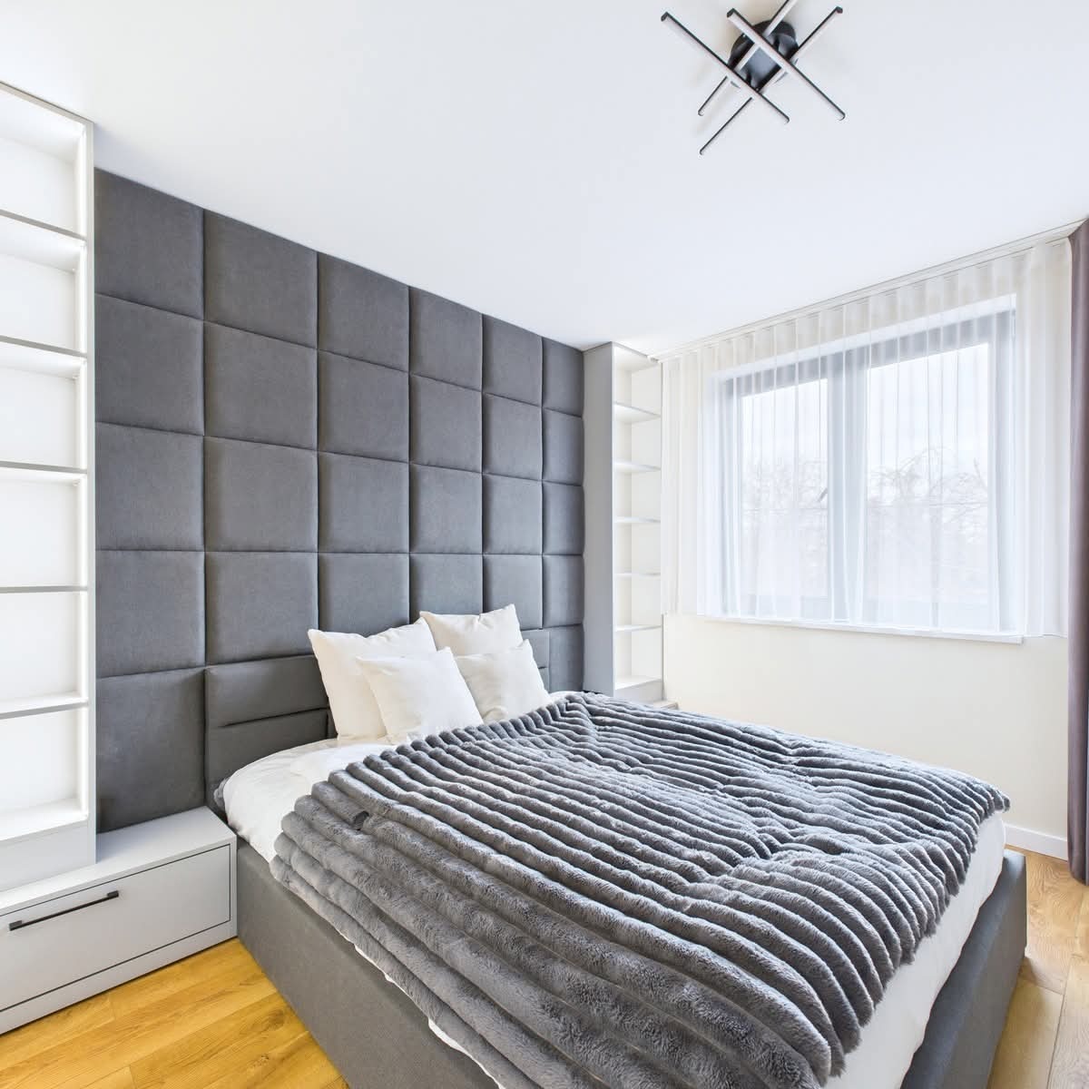 Nowoczesna sypialnia z szarą tapicerowaną ścianą za łóżkiem, białe półki w zabudowie i drewniana podłoga. Minimalistyczny design i stonowana kolorystyka.