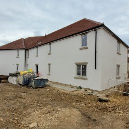 PLZ Painting And Decorating LTD - Biała elewacja budynku wielorodzinnego z czerwonym dachem po malowaniu, widoczne ślady prac wykończeniowych i pojemniki na odpady budowlane.