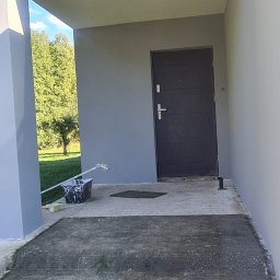 PLZ Painting And Decorating LTD - Świeżo pomalowane wejście do domu w szarym kolorze. Widoczny wałek i kuweta z farbą. Na zewnątrz zieleń drzew i trawnik. Elewacja po renowacji.