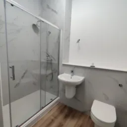 Wykończona łazienka z kabiną prysznicową, umywalką i toaletą. Białe płytki imitujące marmur na ścianach, drewnopodobne panele na podłodze. Minimalistyczny design.