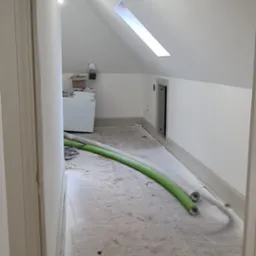 Poddasze po remoncie: białe ściany i sufit ze świetlikiem, szara podłoga, widoczny sprzęt i rury wentylacyjne. Ujęcie zza framugi drzwi.