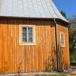 PLZ Painting And Decorating LTD - Drewniana elewacja kościoła z oknami i rynnami w słoneczny dzień. Widoczne detale drewnianej konstrukcji i metalowego dachu. Tło stanowi zieleń drzew i błękitne niebo.