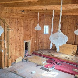 PLZ Painting And Decorating LTD - Wnętrze drewnianego pomieszczenia w trakcie malowania. Widoczne rusztowanie, zabezpieczone lampy i ściany pokryte świeżą warstwą bejcy. Częściowo odsłonięta podłoga.