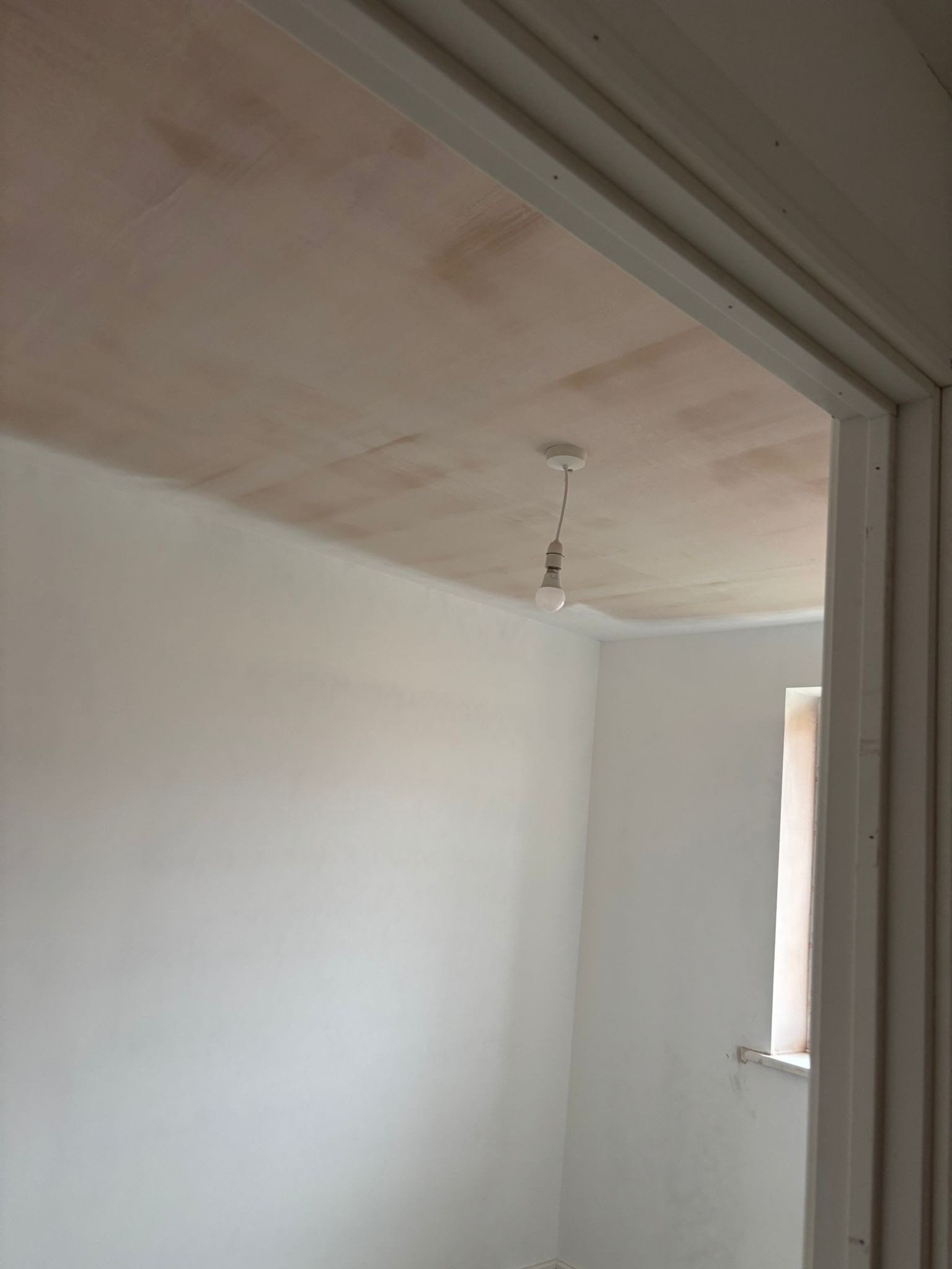 Pokój po szpachlowaniu, przygotowany do malowania. Widoczne ślady po szpachli na suficie i ścianach. Żarówka wisi na kablu. Biała futryna drzwiowa.