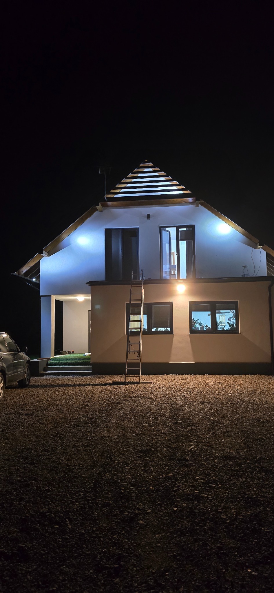 Oświetlona elewacja domu po renowacji w Płoniawach-Bramurze, z drabiną opartą o ścianę. Widoczne okna i elementy wykończeniowe dachu.