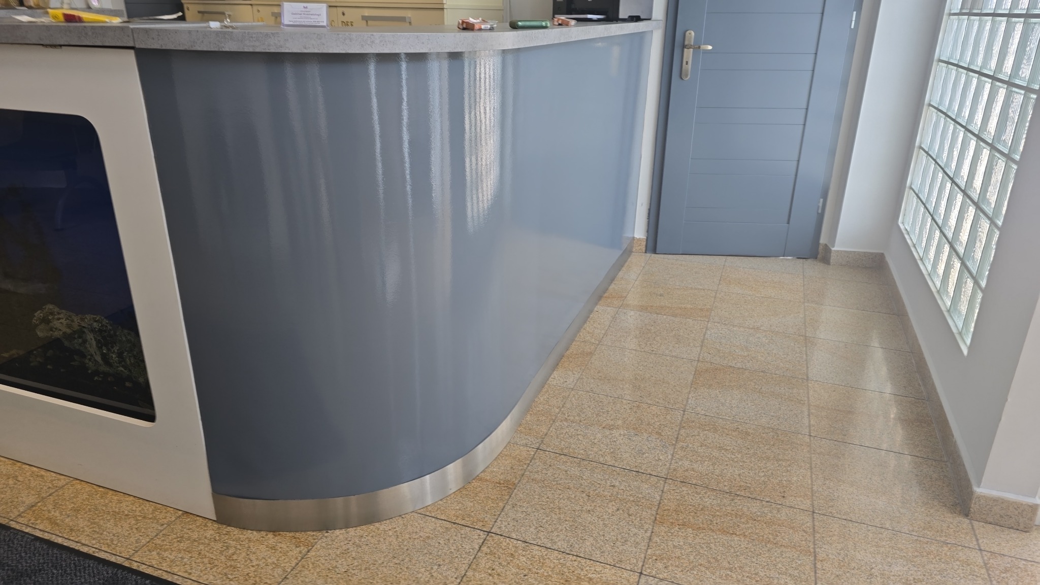 Gładka, szara lada recepcyjna z akwarium, świeżo po malowaniu, z widocznym połyskiem farby. W tle szare drzwi i ściana z luksferami.