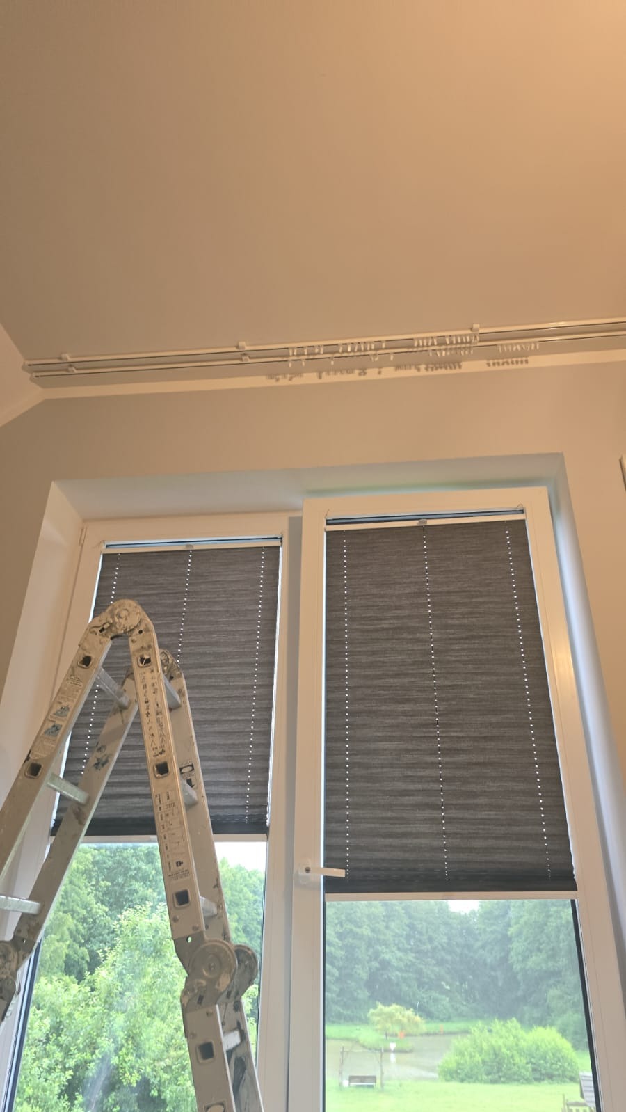 Białe okno z szarymi plisami, drabina oparta o ramę, widok na zieleń za oknem. Detal wnętrza po montażu plis okiennych, widoczne prowadnice sufitowe.