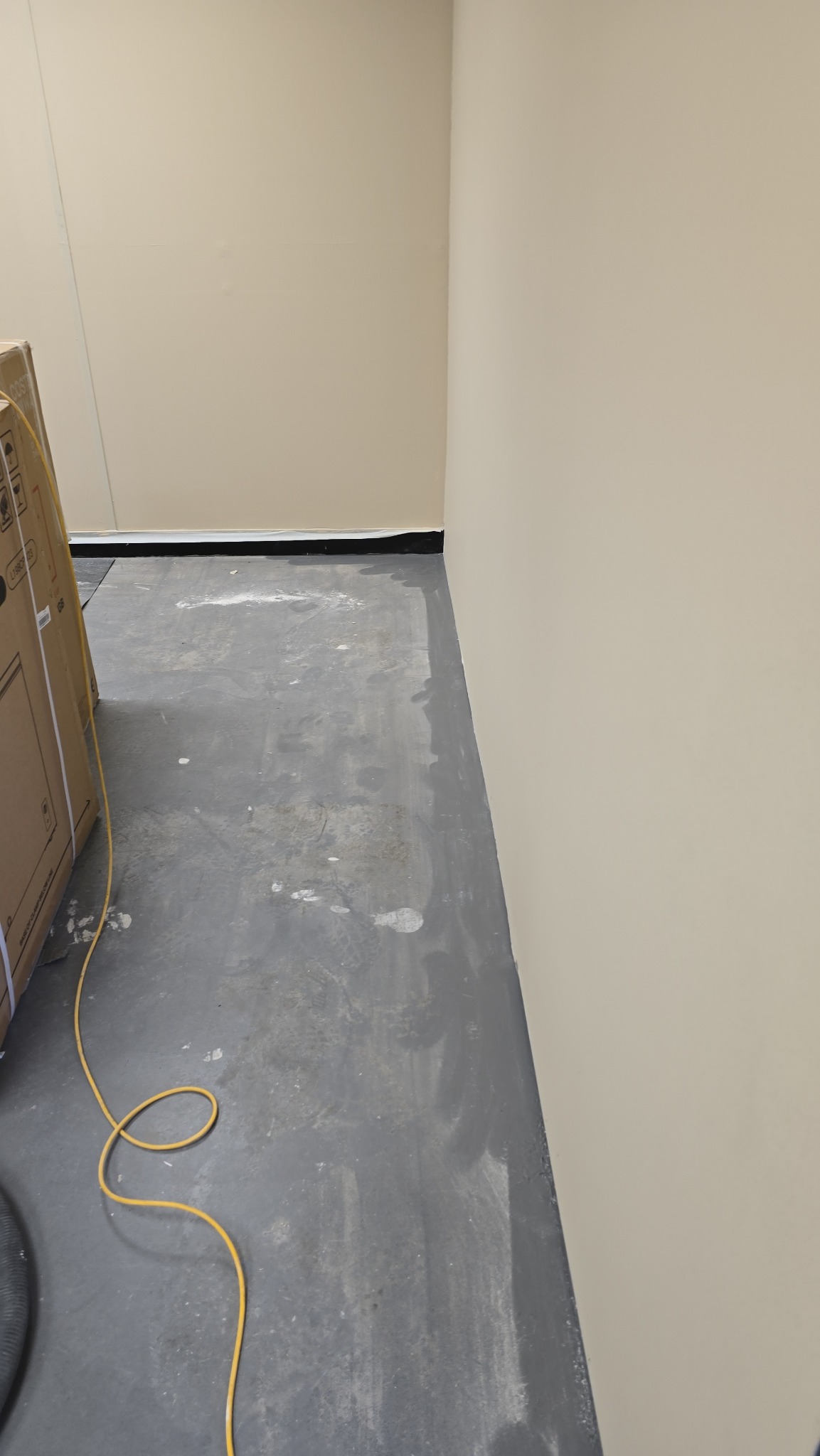 Świeżo pomalowana ściana w jasnym odcieniu beżu, kontrastująca z szarą podłogą zabezpieczoną folią malarską. Widoczny kabel i karton, co sugeruje trwające prace wykończeniowe.