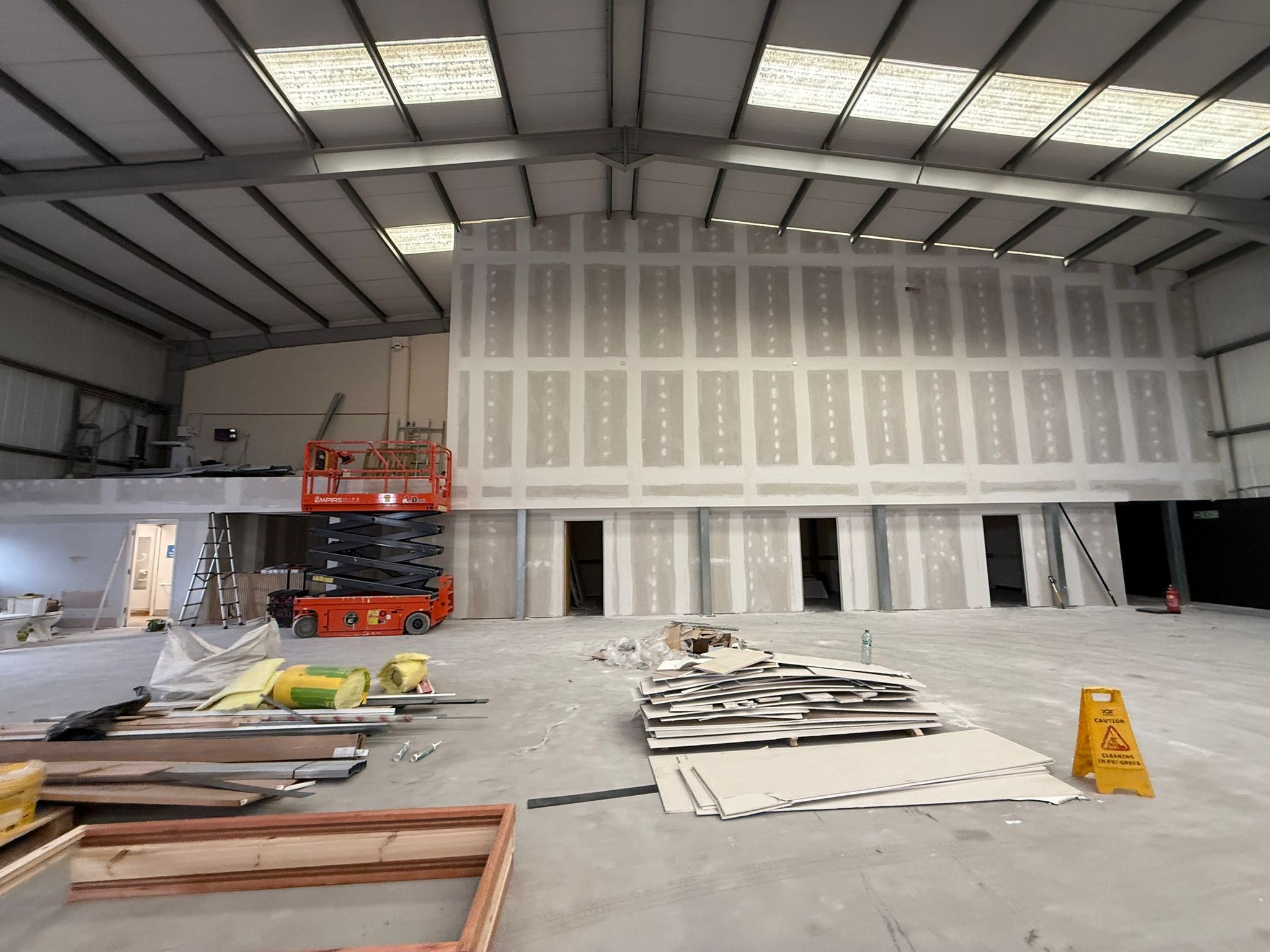Wnętrze hali w trakcie prac wykończeniowych: ściany z płyt kartonowo-gipsowych, podnośnik nożycowy, materiały budowlane i znak ostrzegawczy na posadzce.