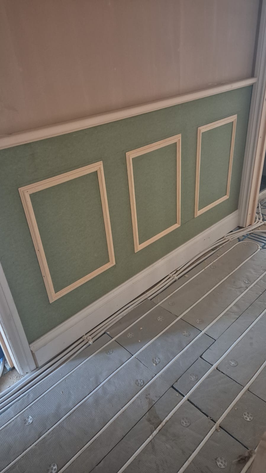 Ściana z dekoracyjnymi ramkami i przygotowanym podłożem pod malowanie, widoczne elementy instalacji grzewczej w podłodze. Prace wykończeniowe w pomieszczeniu.