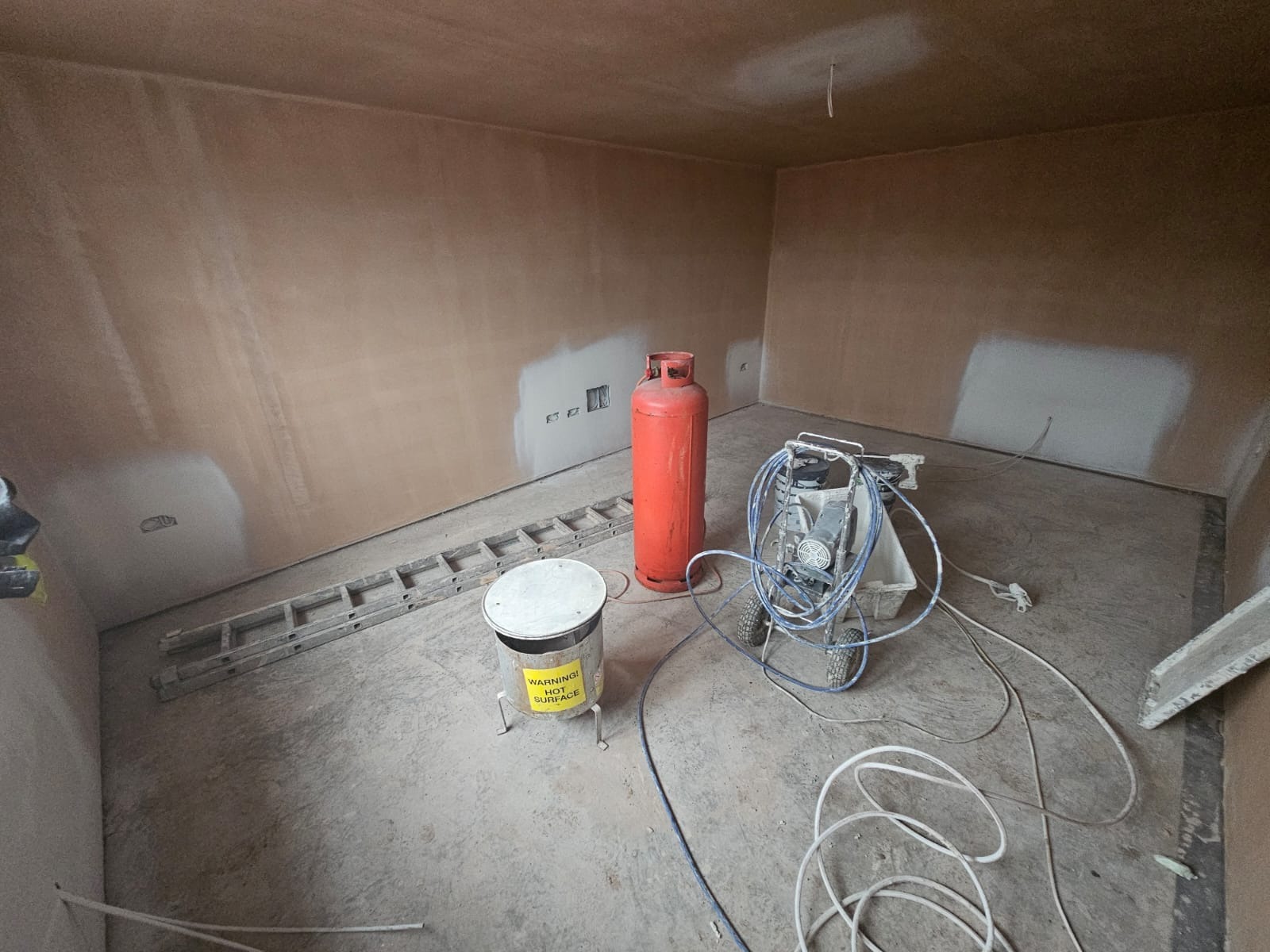 Pokój po gładzi, przygotowany do malowania natryskowego. Widoczny agregat malarski, butla gazowa i drabina. Ściany częściowo zagruntowane.