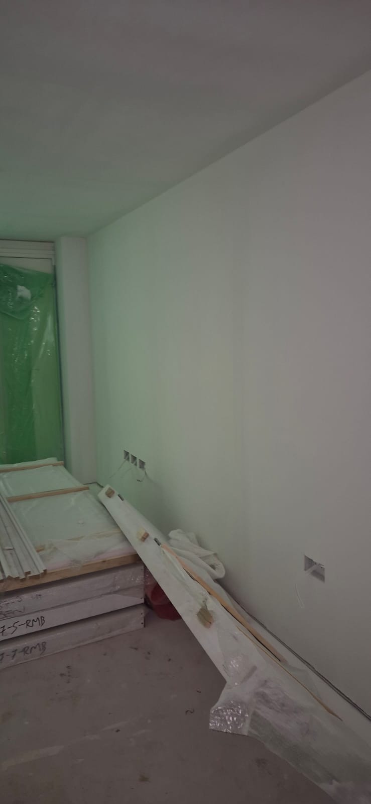 Świeżo pomalowane na biało wnętrze pokoju z widocznymi puszkami elektrycznymi i materiałami budowlanymi pod ścianą, zabezpieczonymi folią.