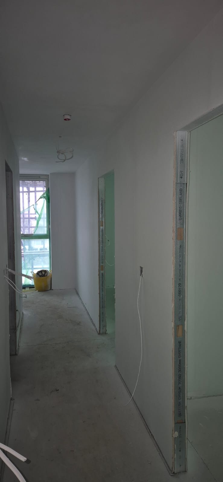 Korytarz w trakcie malowania ścian na biało, widoczne profile gipsowo-kartonowe przy drzwiach i wystające kable. Żółte wiadro w tle.