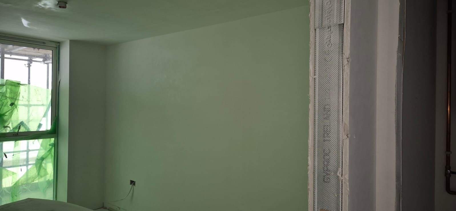 Świeżo pomalowane wnętrze pokoju na jasnozielono, z oknem zabezpieczonym folią i fragmentem konstrukcji ścianki działowej z płyty gipsowej po prawej stronie.