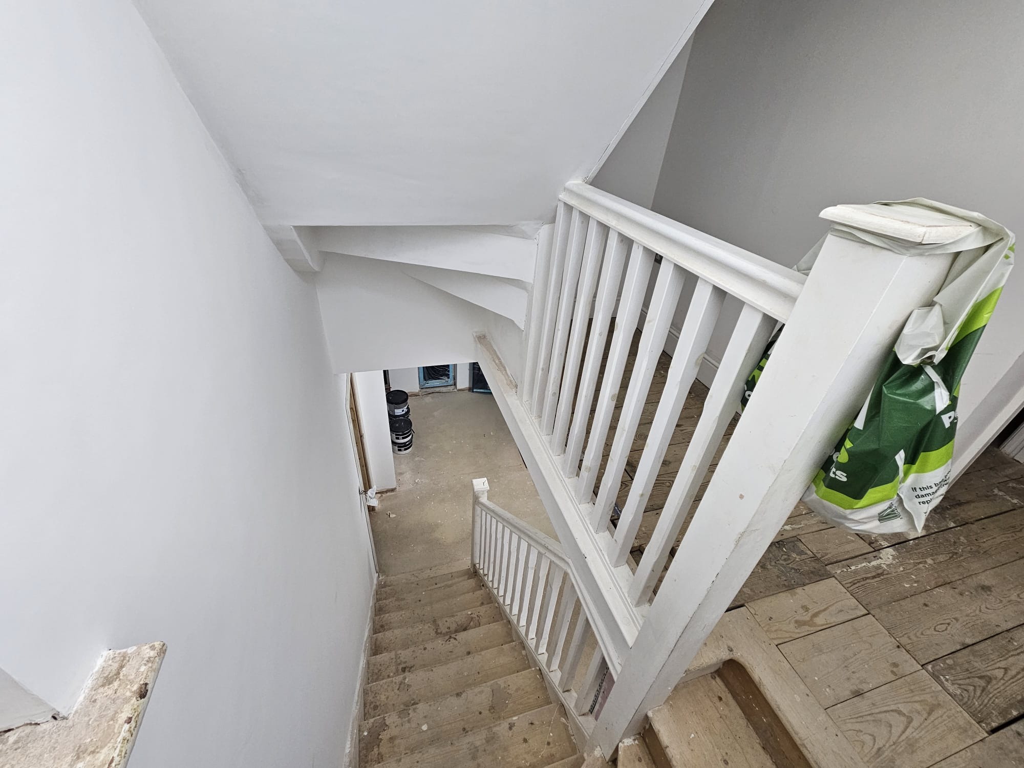 Białe schody po malowaniu, widok z góry. Ściany i sufit świeżo pomalowane na biało, widoczne ślady po remoncie na schodach i podłodze. Torby z farbą opierają się o balustradę.