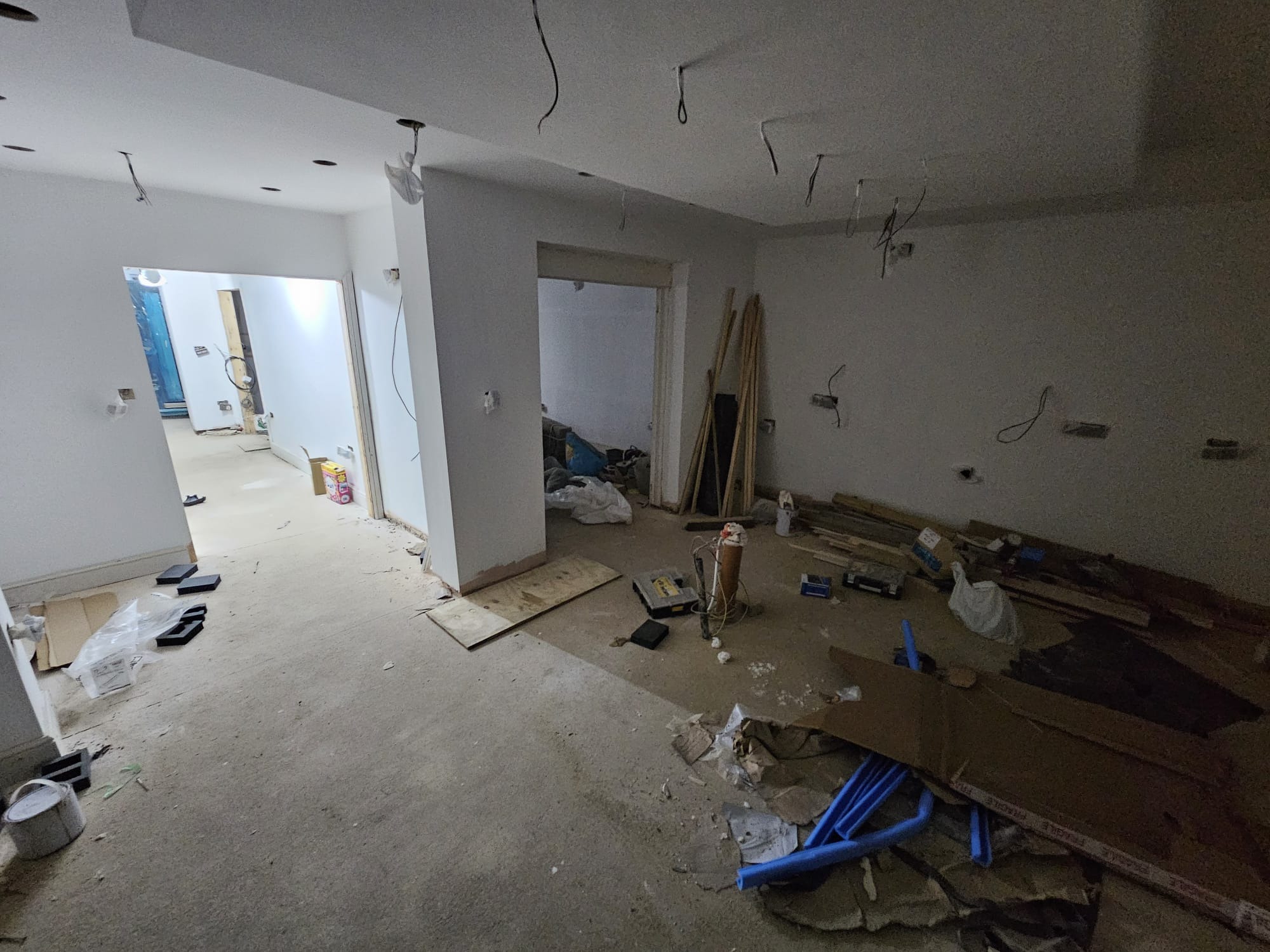 Wnętrze w trakcie malowania: białe ściany, widoczne narzędzia, przewody elektryczne wystające z sufitu. Podłoga zabezpieczona kartonami i folią. Etap prac wykończeniowych.