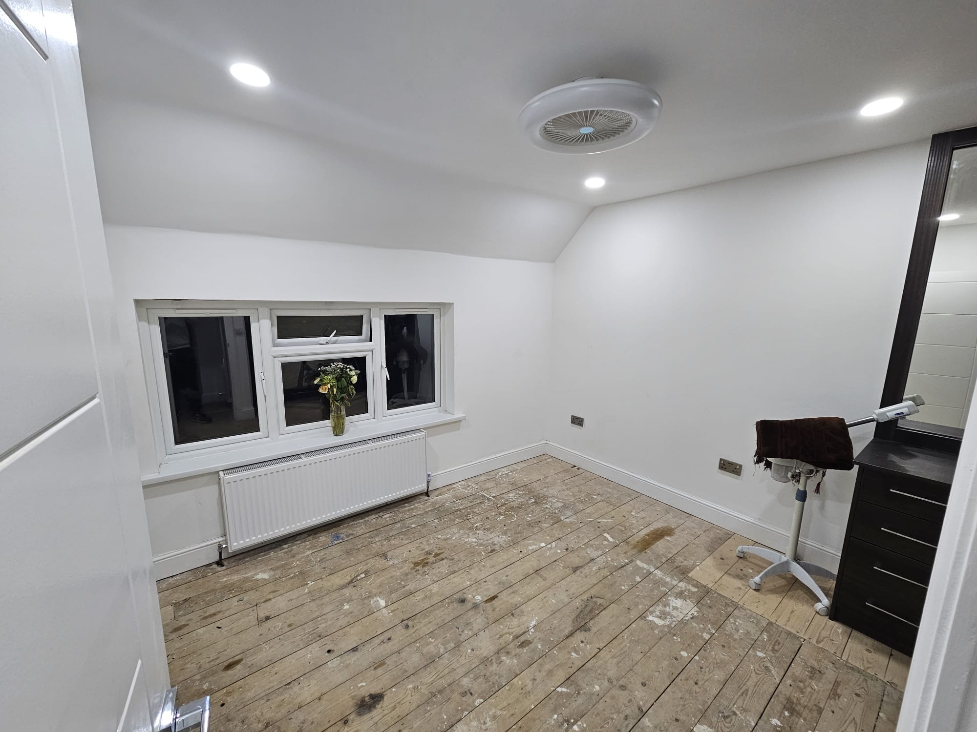 Pomalowane na biało ściany pokoju z drewnianą podłogą przed wykończeniem, widoczne okno z kwiatami, grzejnik i toaletka z lustrem. Świeżo po malowaniu.