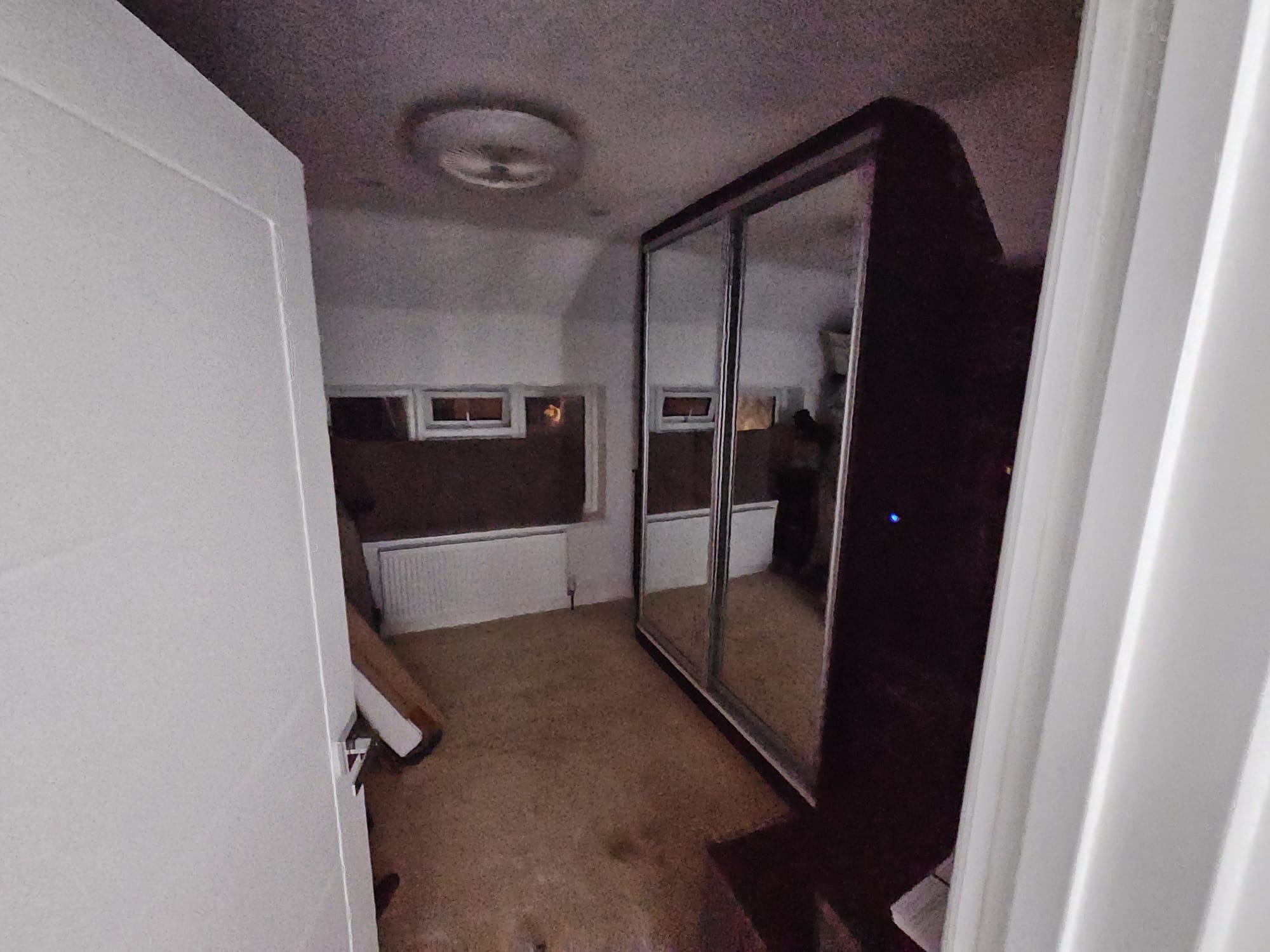 Wnętrze pokoju z brązową podłogą, białymi ścianami i grzejnikiem. Widoczna szafa z lustrem i białe drzwi. Sufit z okrągłą lampą.