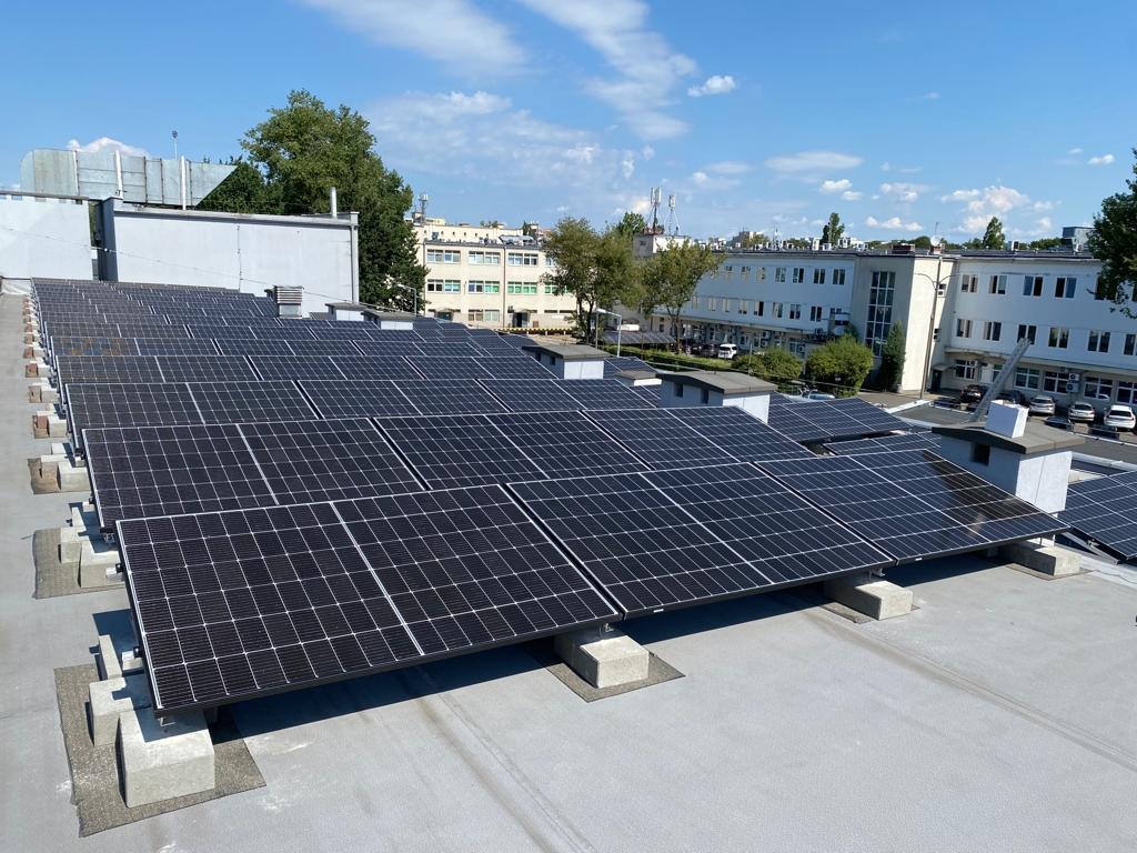 Instalacja fotowoltaiczna na dachu budynku z widokiem na miejski krajobraz w słoneczny dzień. Panele ustawione w rzędach, betonowe podstawy, jasne niebo z chmurami.