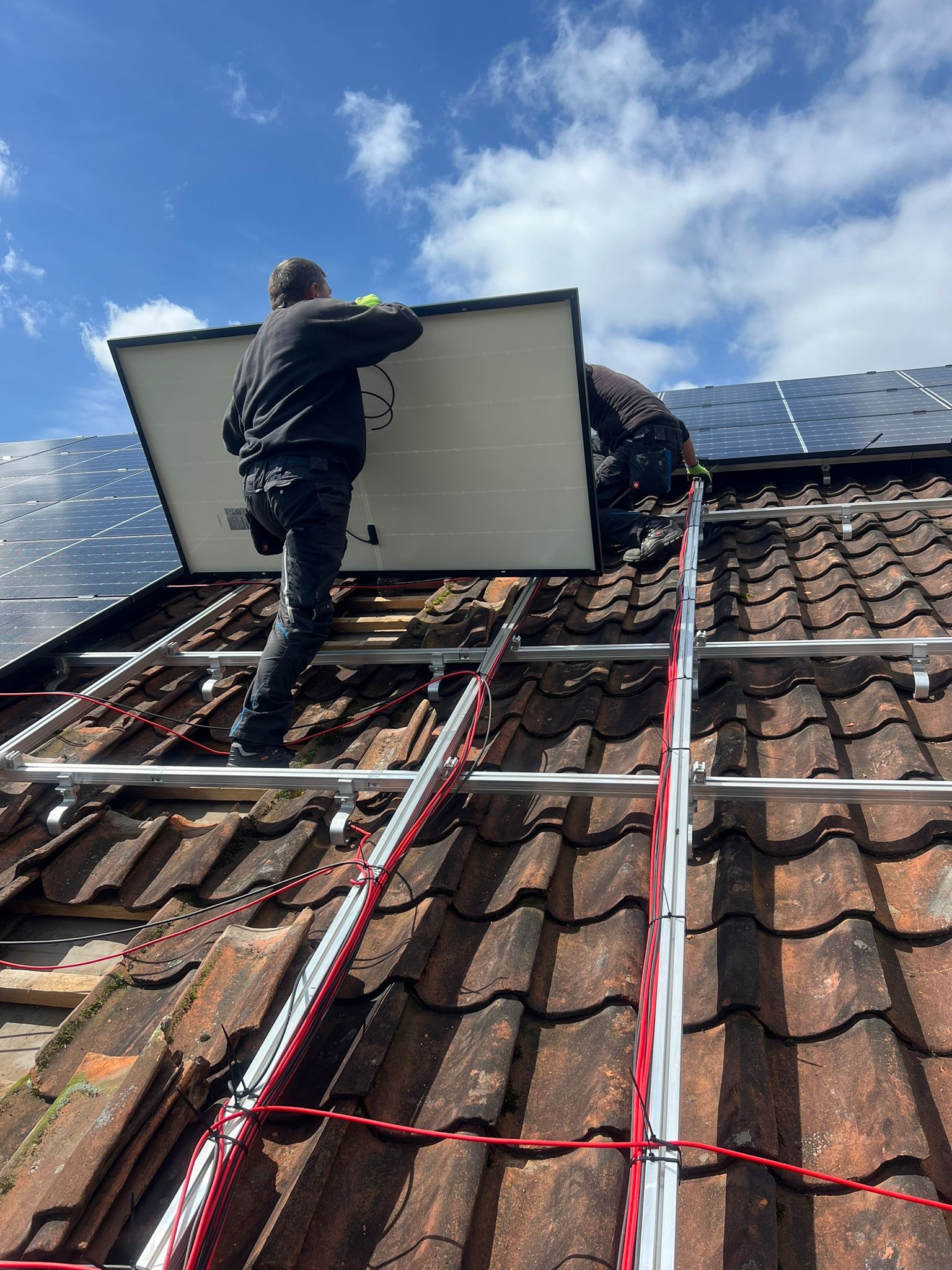Montaż paneli fotowoltaicznych na dachu z czerwoną dachówką. Widoczne aluminiowe profile, kable i dwóch monterów instalujących panel. Błękitne niebo w tle.