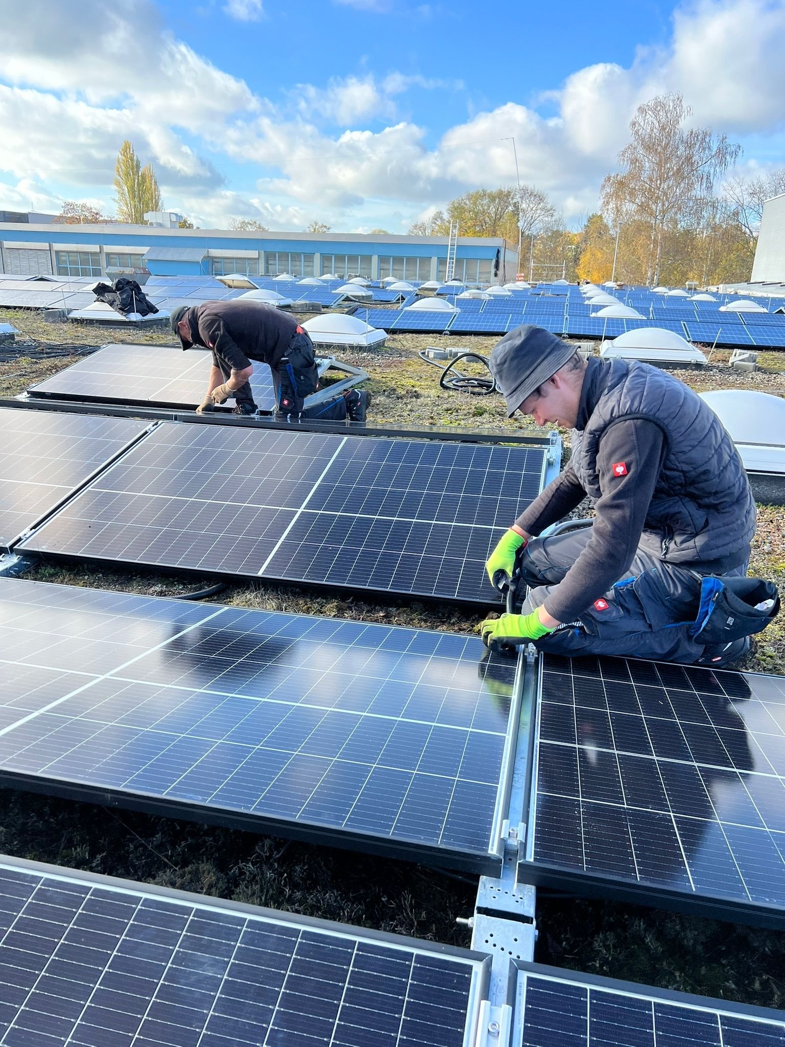 Instalatorzy w kombinezonach montują panele fotowoltaiczne na dachu budynku w słoneczny dzień. Widoczne narzędzia i elementy konstrukcyjne instalacji.