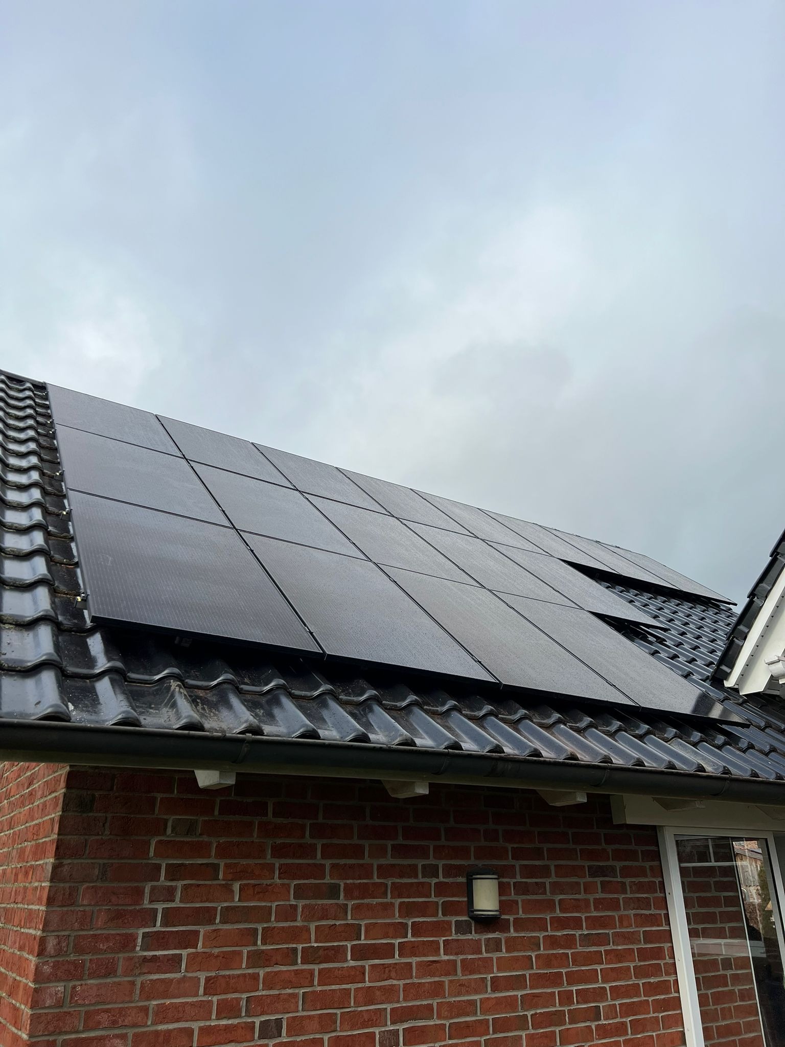 Ciemne panele fotowoltaiczne na czarnym, błyszczącym dachu z dachówki, widoczne z dołu, ceglana ściana budynku w tle, pochmurne niebo.