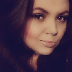 Portret kobiety z długimi, brązowymi włosami, delikatnym makijażem i błyszczącymi ustami, uchwycony w miękkim, rozproszonym świetle.