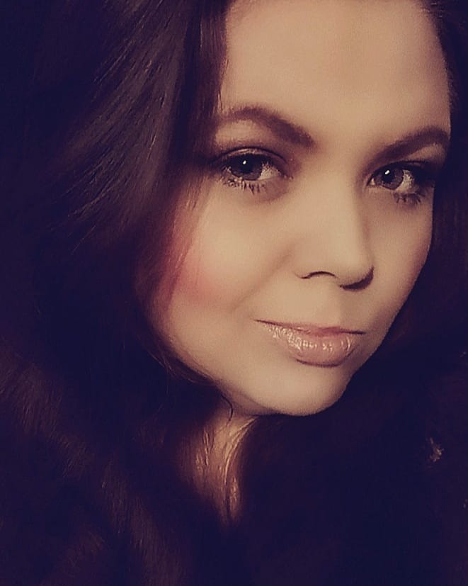 Portret kobiety z długimi, brązowymi włosami, delikatnym makijażem i błyszczącymi ustami, uchwycony w miękkim, rozproszonym świetle.