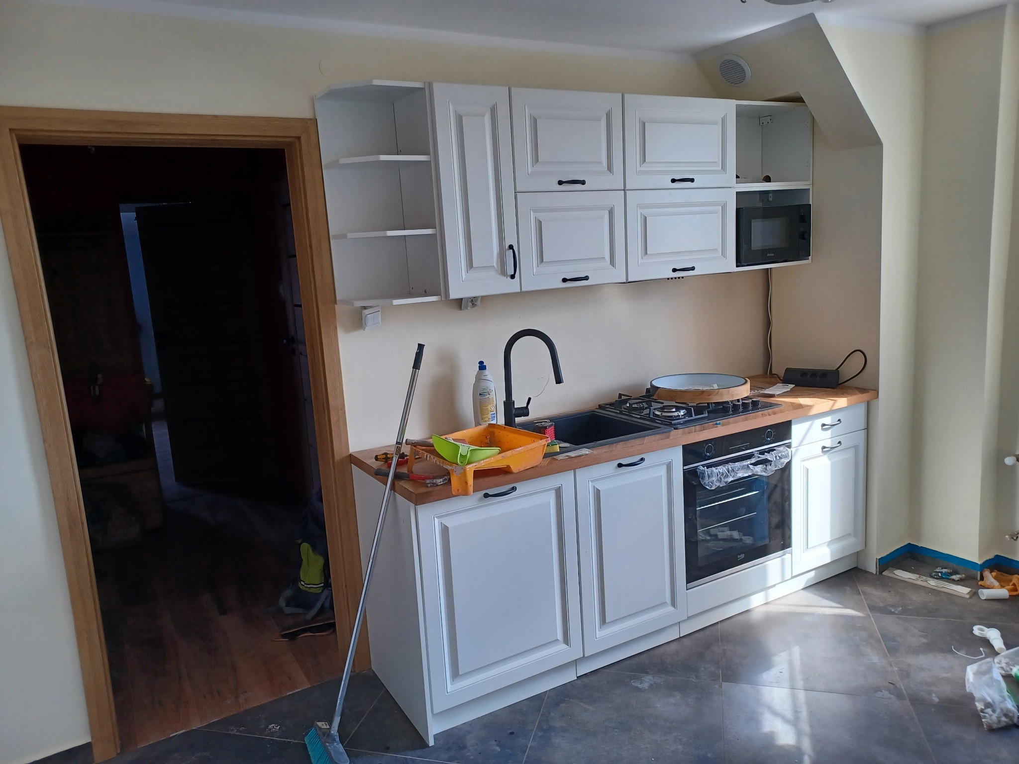 Biała kuchnia STILO z drewnianym blatem, czarnym zlewem i kuchenką. Widoczne narzędzia malarskie sugerują świeży remont. Ustawiona w pomieszczeniu ze skośnym sufitem.