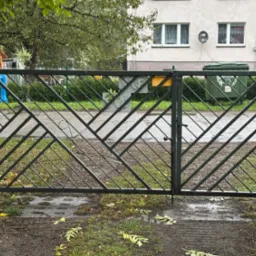 Nowoczesna, dwuskrzydłowa brama wjazdowa z metalu, z ukośnym wzorem kratownicy. Szara kolorystyka bramy komponuje się z otoczeniem, w tle budynek mieszkalny i zieleń.