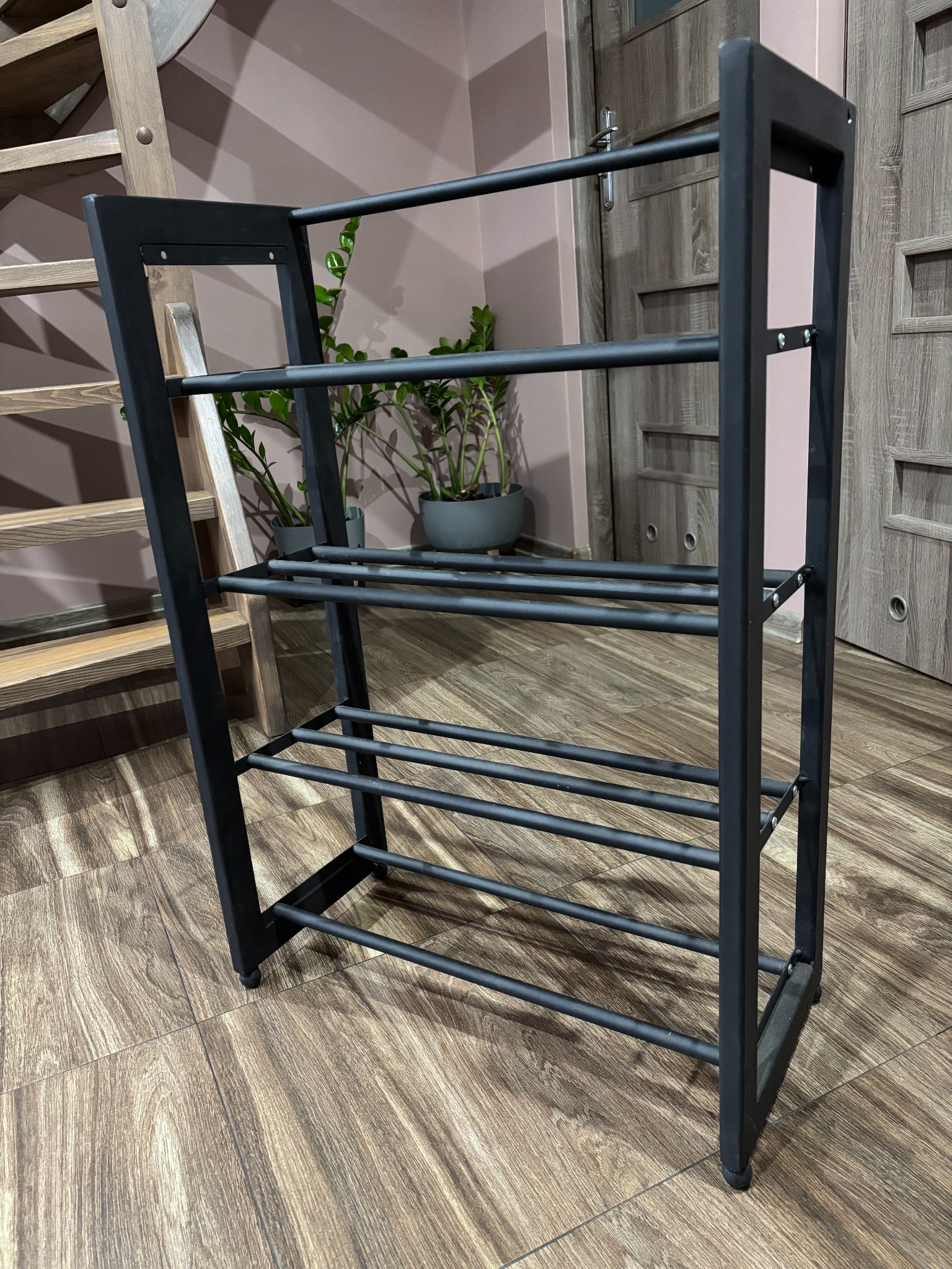 Czarny, metalowy regał na buty z trzema poziomami, widoczny na tle drewnianej podłogi i ściany w odcieniach beżu. W tle fragment drewnianych schodów i doniczka z rośliną.