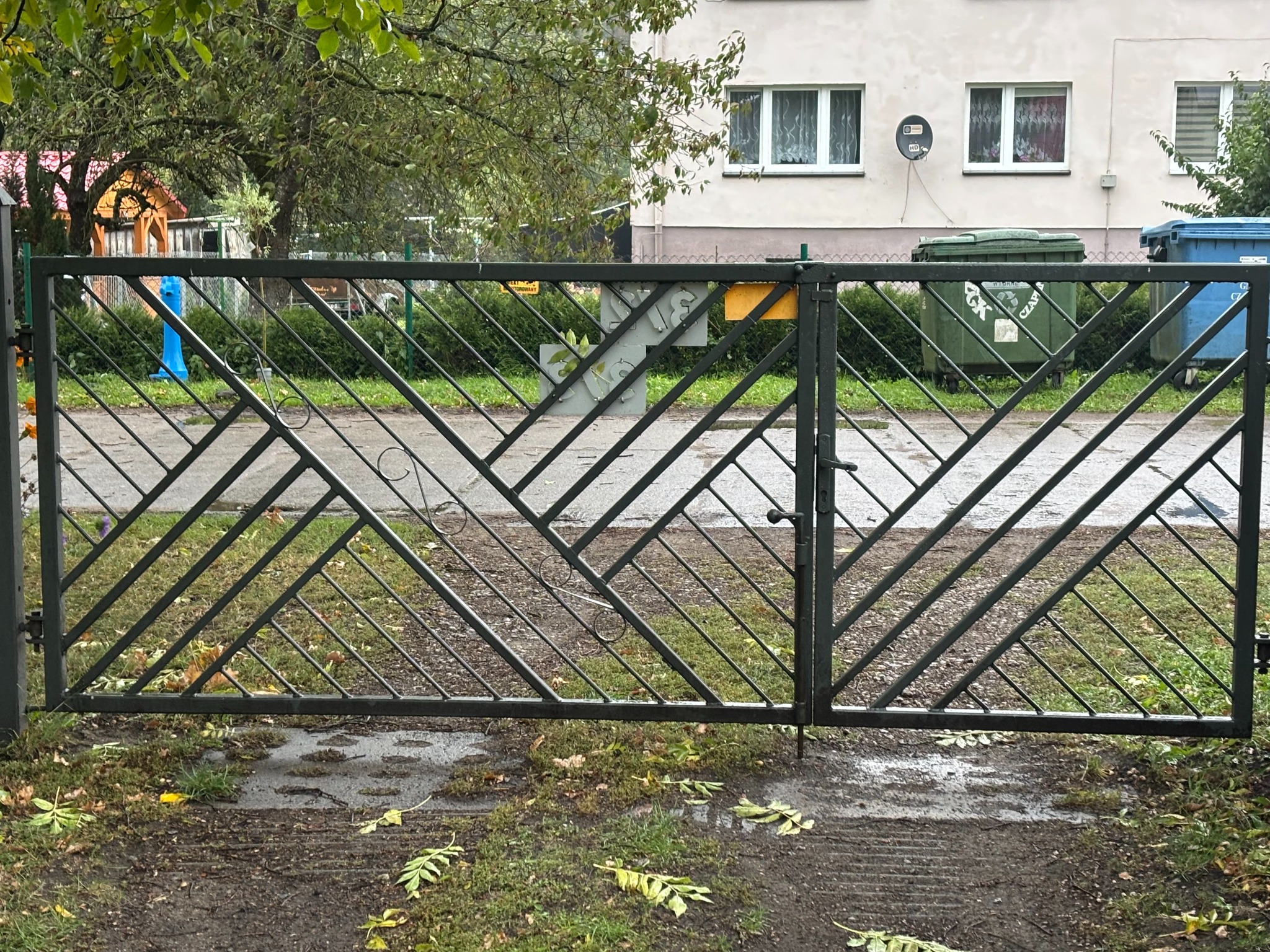 Nowoczesna, dwuskrzydłowa brama wjazdowa z metalu, z ukośnym wzorem kratownicy. Szara kolorystyka bramy komponuje się z otoczeniem, w tle budynek mieszkalny i zieleń.