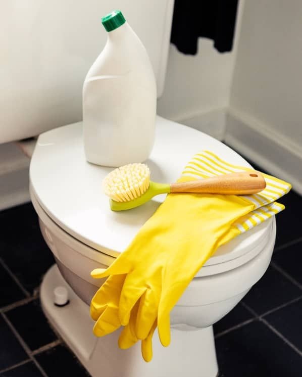 Środki czystości na desce sedesowej: szczotka z zielonym uchwytem, żółte rękawice gumowe, biała butelka z zieloną nakrętką i ściereczka w żółte paski.