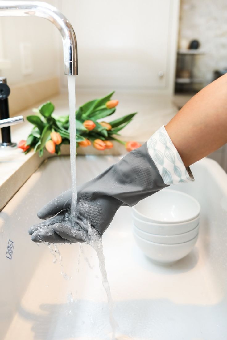 Ręka w szarej rękawicy ochronnej pod strumieniem wody w zlewie, obok leżą tulipany i miseczki. Domowe porządki w kuchni.