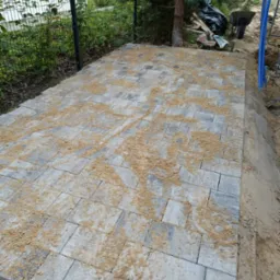 Układanie szarej kostki brukowej na podsypce piaskowej w ogrodzie, widoczne narzędzia i fragment ogrodzenia.