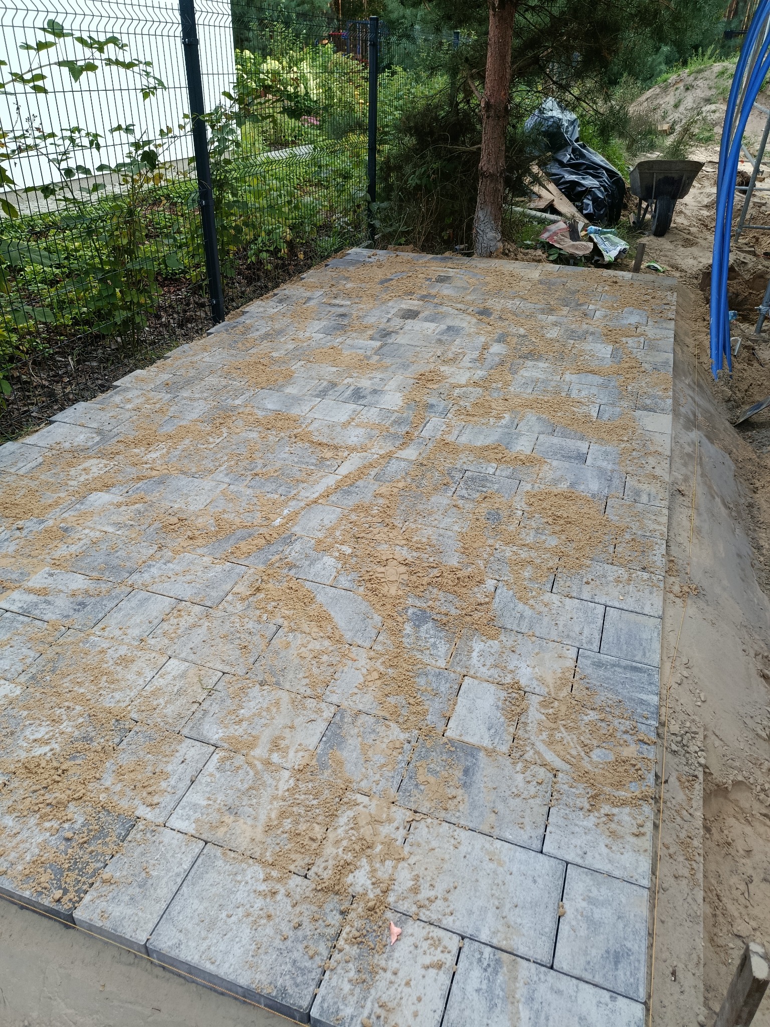 Układanie szarej kostki brukowej na podsypce piaskowej w ogrodzie, widoczne narzędzia i fragment ogrodzenia.