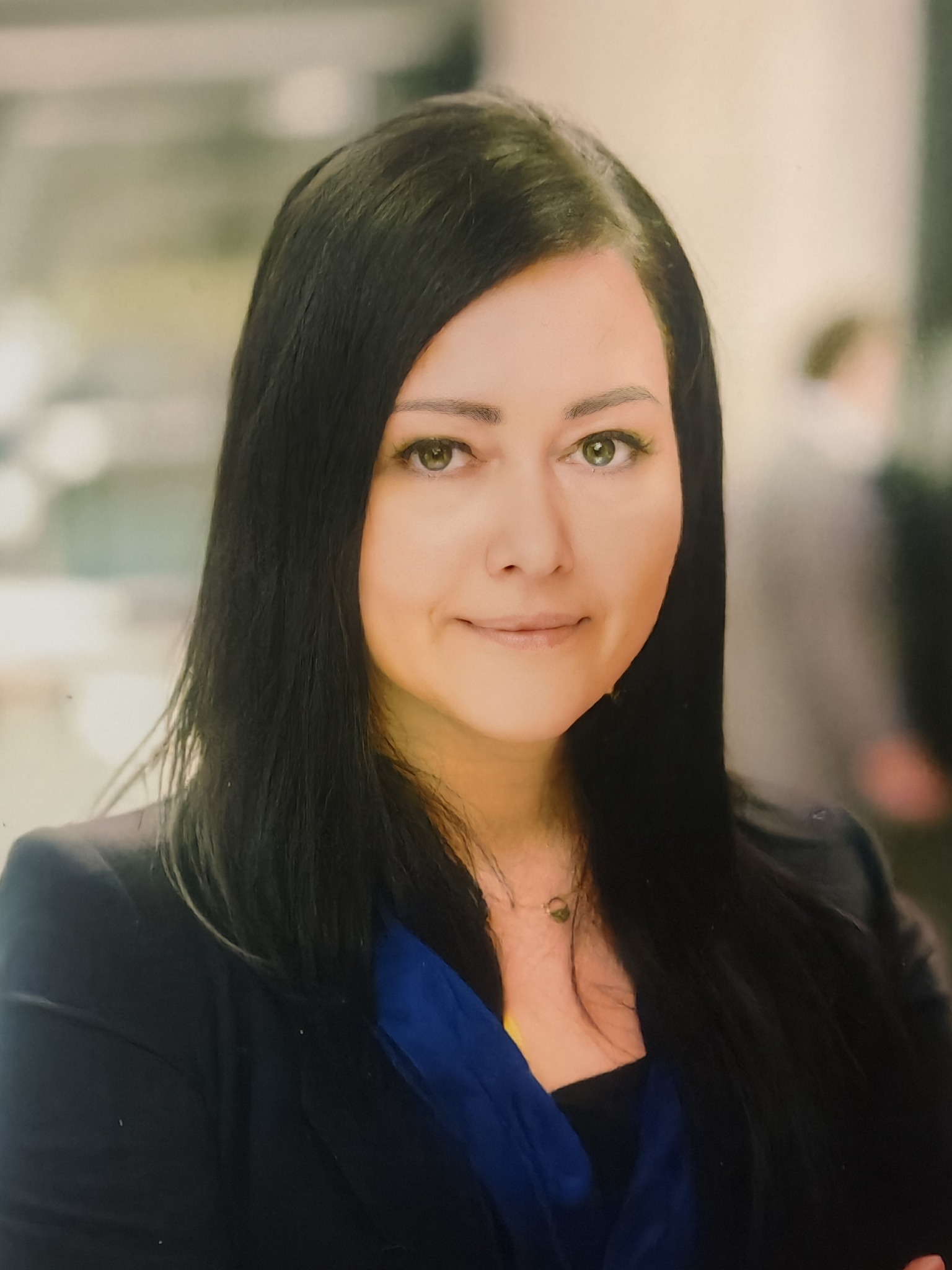 Portret kobiety z długimi, czarnymi włosami, ubranej w niebieską bluzkę i czarną marynarkę, z delikatnym uśmiechem, na rozmytym tle biurowym.