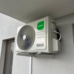 COLDWARM Kacper Rochniak - Zamontowana jednostka zewnętrzna klimatyzacji Kaisai na szarej elewacji budynku. Widoczne okno i fragment dachu. Na klimatyzatorze naklejka z danymi kontaktowymi.