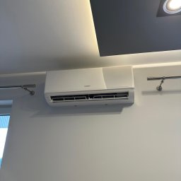 COLDWARM Kacper Rochniak - Biała klimatyzacja Kaisai na jasnej ścianie pod sufitem z oświetleniem. Widoczne uchwyty na ścianie, minimalistyczny design.