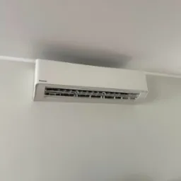 Biała klimatyzacja Panasonic zamontowana na jasnej ścianie w pokoju. Widoczny fragment kosza z wikliny w rogu. Minimalistyczny design wnętrza.