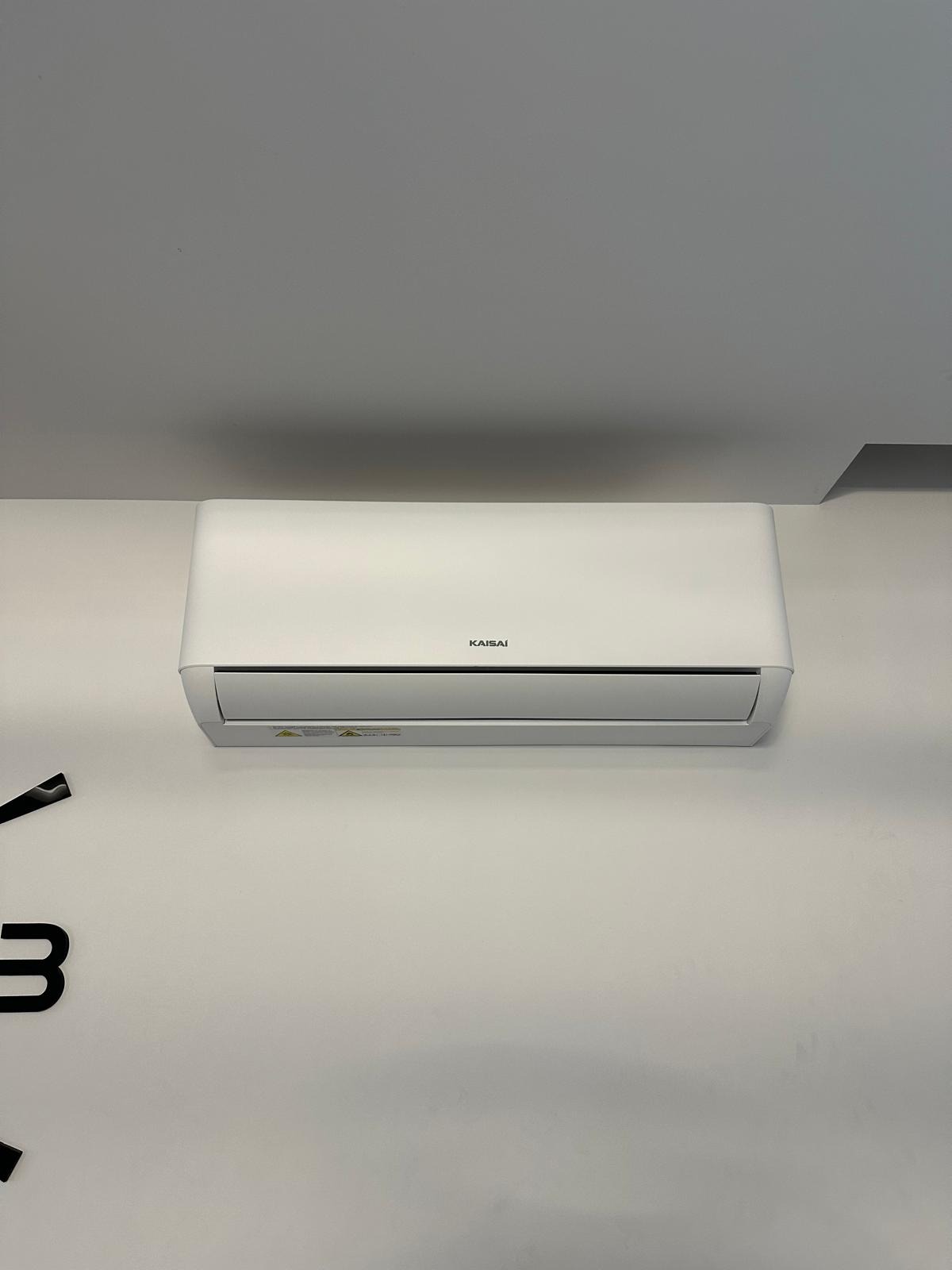 Biała klimatyzacja Kaisai zamontowana na jasnej ścianie, widoczny fragment czarnego zegara po lewej stronie. Minimalistyczny design, czyste linie.