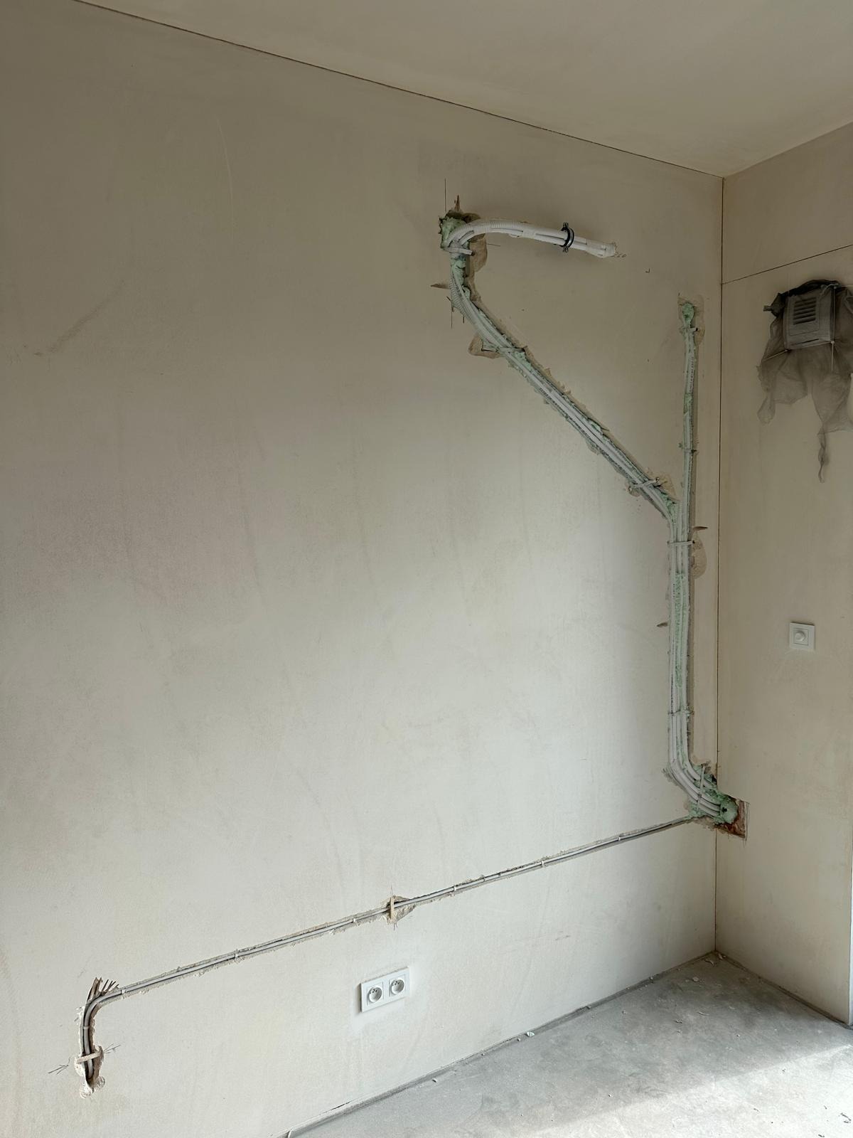 Białe rury wentylacyjne i kable elektryczne zamontowane na surowej ścianie w rogu pokoju, widoczne puszki elektryczne i kratka wentylacyjna.