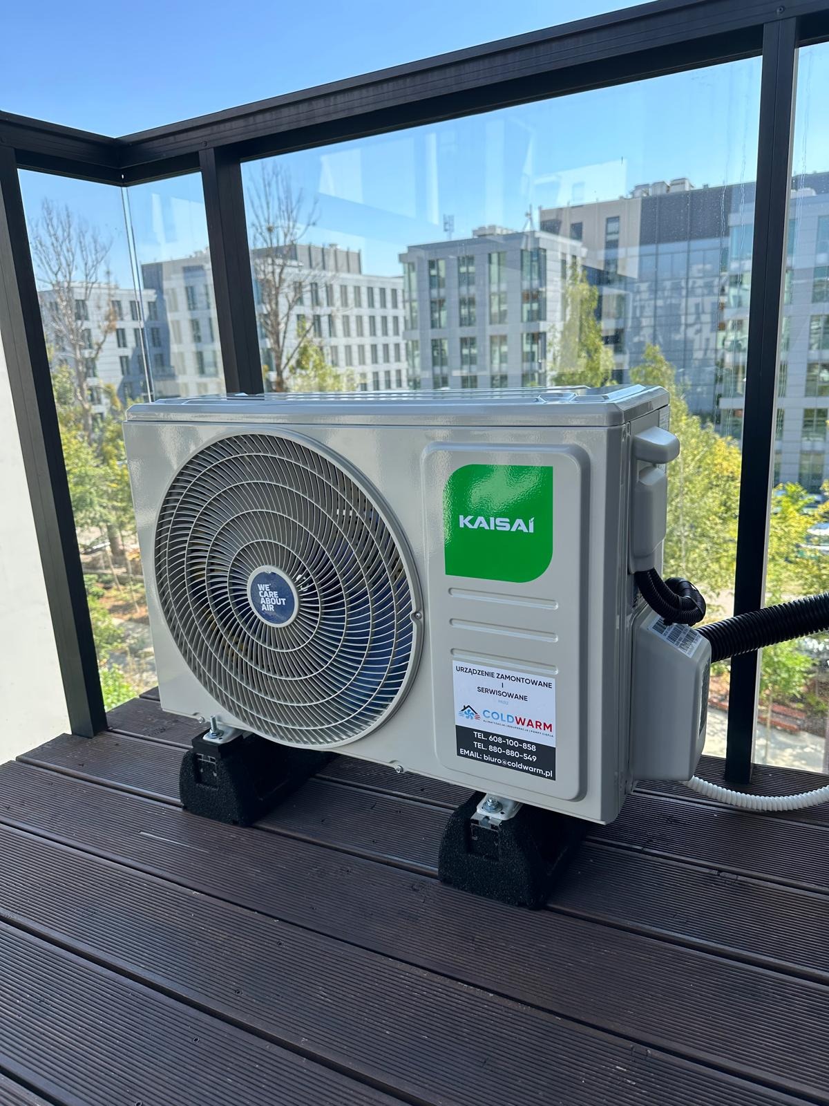 Zamontowana klimatyzacja Kaisai na balkonie w Warszawie, widok na nowoczesne budynki. Urządzenie stoi na gumowych podkładkach. Widoczna tabliczka firmy Coldwarm.