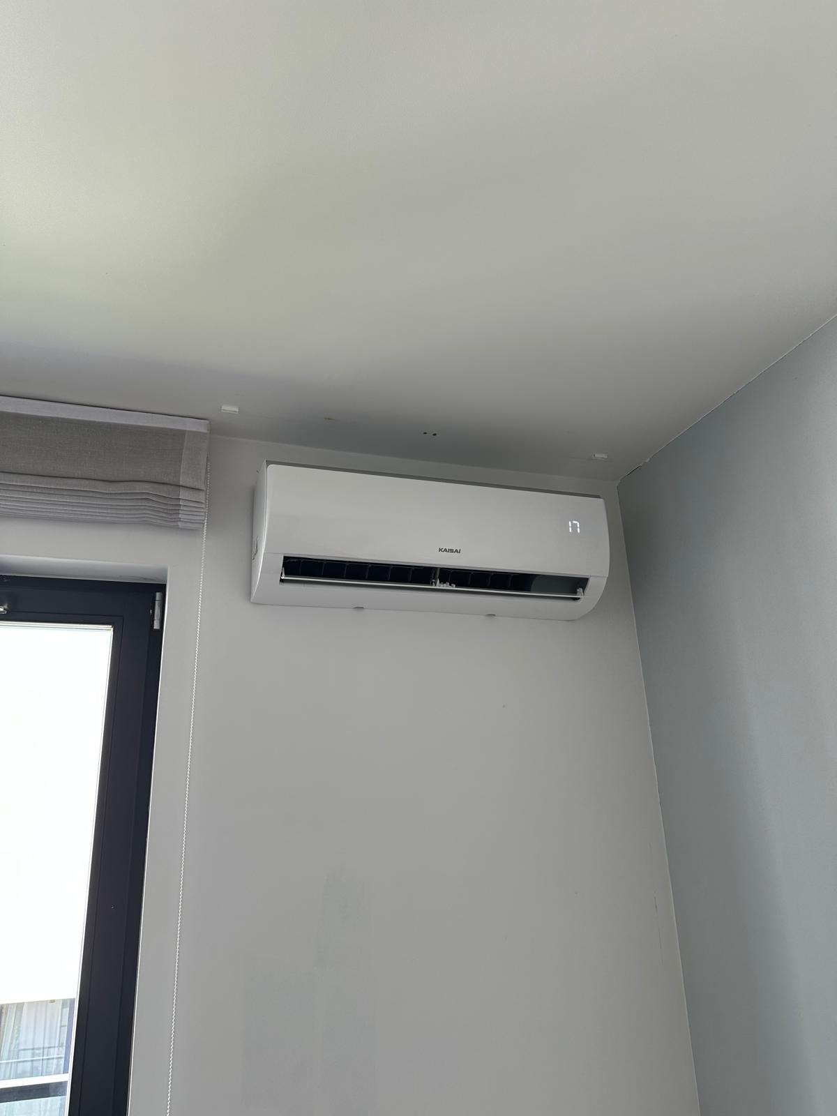 Biały klimatyzator Kaisai, zamontowany na szarej ścianie w pokoju z roletami. Na wyświetlaczu widoczna temperatura 17 stopni.