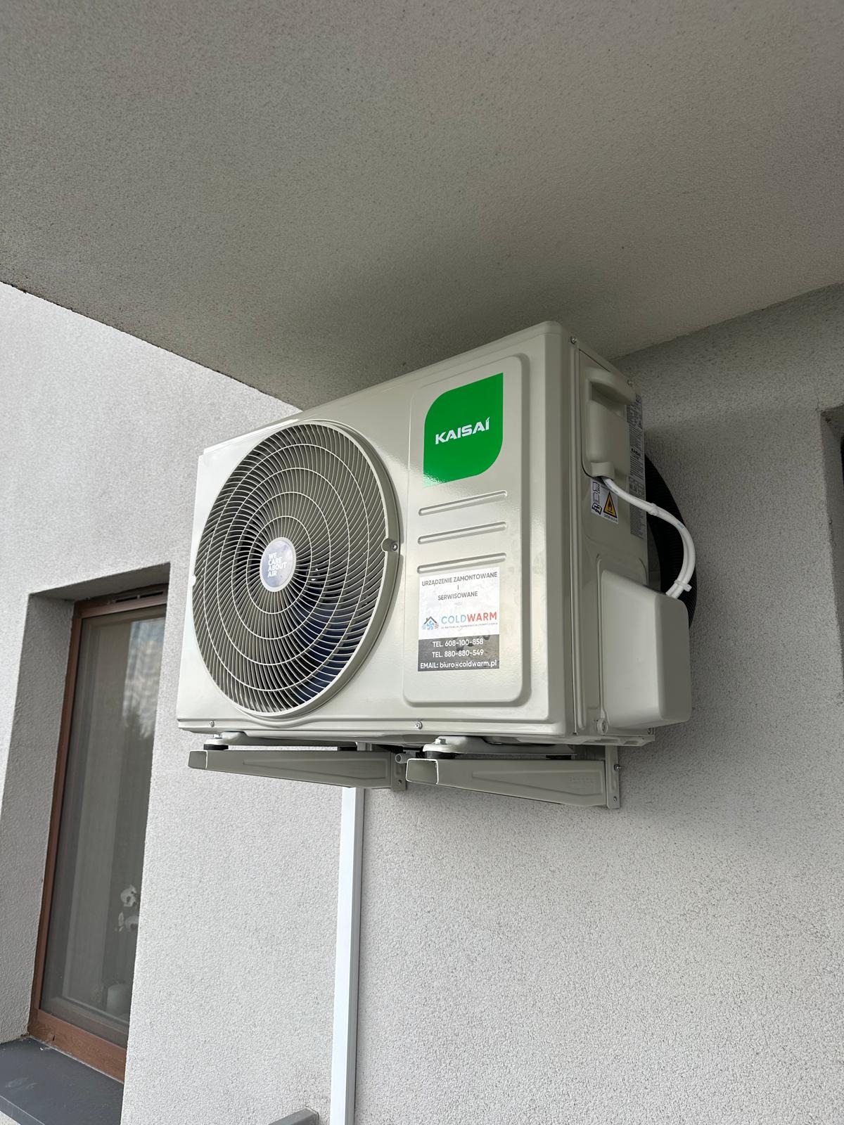 Zamontowana jednostka zewnętrzna klimatyzacji Kaisai na szarej elewacji budynku. Widoczne okno i fragment dachu. Na klimatyzatorze naklejka z danymi kontaktowymi.
