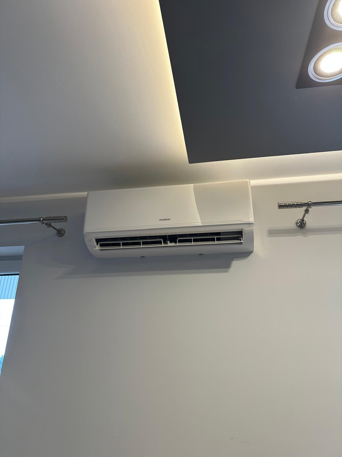 Biała klimatyzacja Kaisai na jasnej ścianie pod sufitem z oświetleniem. Widoczne uchwyty na ścianie, minimalistyczny design.