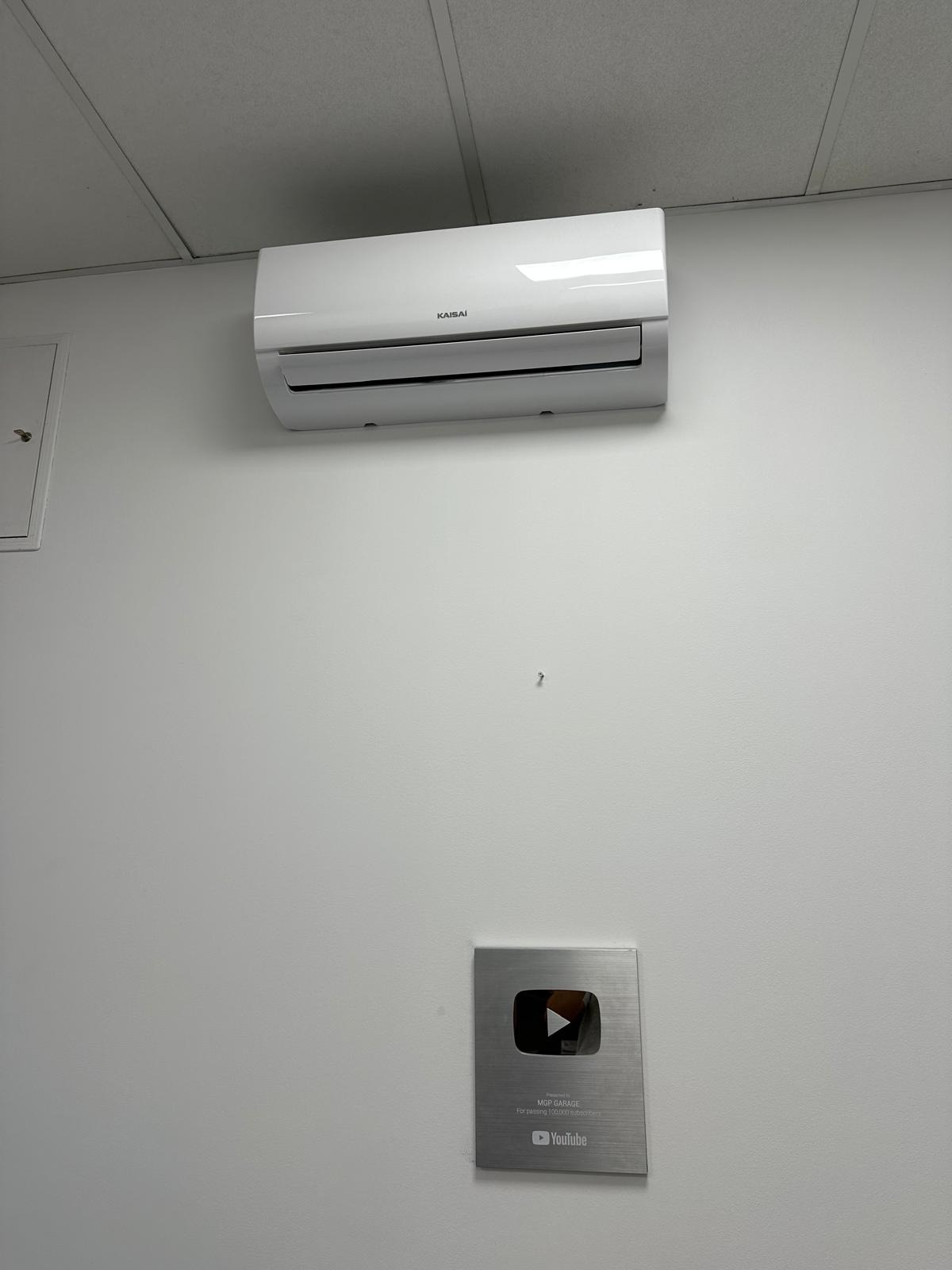 Biały klimatyzator Kaisai zamontowany na ścianie nad srebrną nagrodą YouTube dla kanału MGP Garage, w jasnym pomieszczeniu z kasetonowym sufitem.