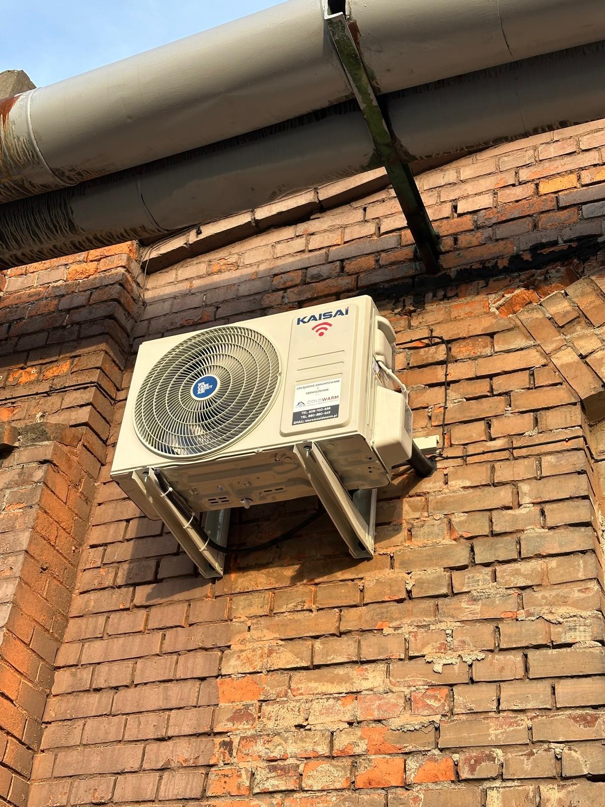 Klimatyzator Kaisai zamontowany na ceglanej ścianie budynku. Widoczne rury instalacyjne i metalowe wsporniki. Ujęcie z dołu, podkreślające solidność montażu.