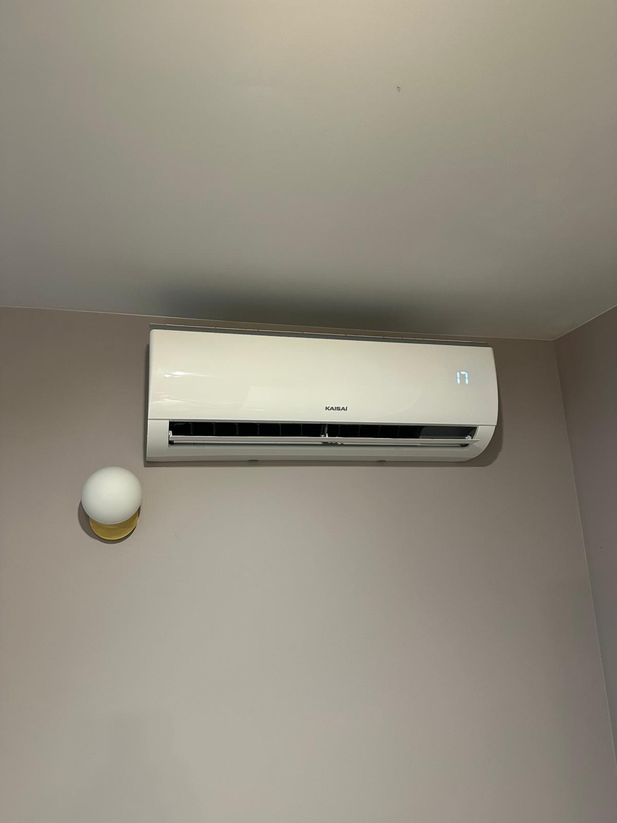 Biała klimatyzacja Kaisai na ścianie w odcieniach beżu, obok lampka nocna. Na wyświetlaczu urządzenia widoczna temperatura 17 stopni.